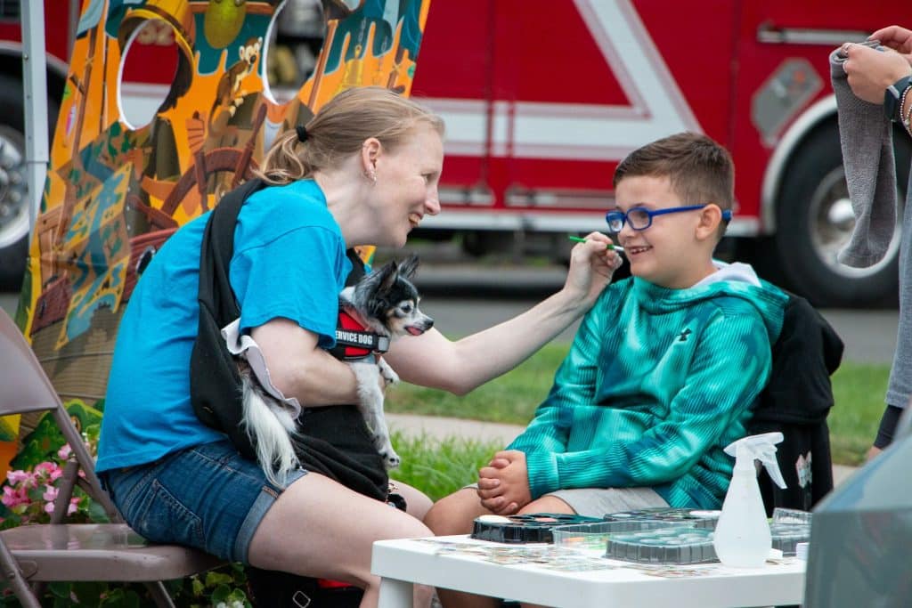 A boy gets his face painted by a member of the Enfield Youth Council, accompanied by a service dog. 